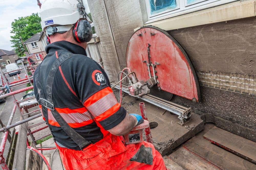 Construction worker cutting exterior wall with power saw on scaffolding in residential area.