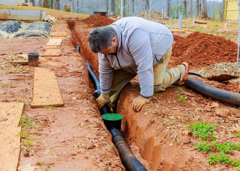 Person installing drainage pipe in a dug trench on a construction site outdoors.